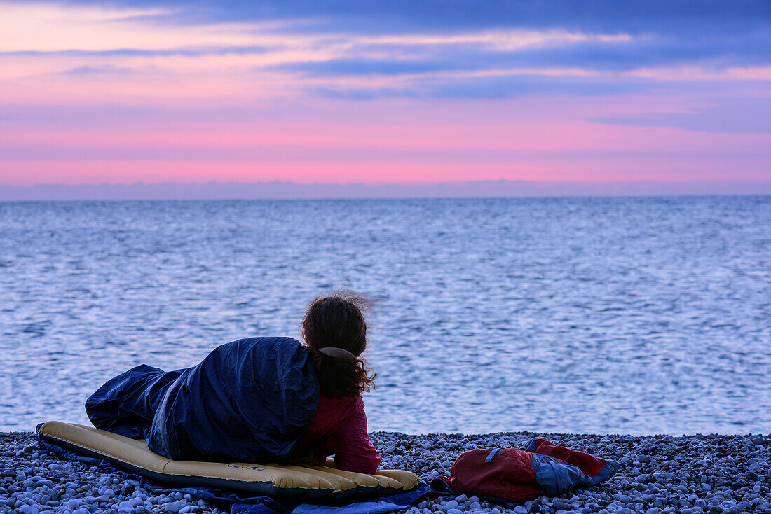 Woman laying in sleeping bag at beach and looking at Mediterranean, Selvaggio Blu, National Park of the Bay of Orosei and Gennargentu, Sardinia, Italy