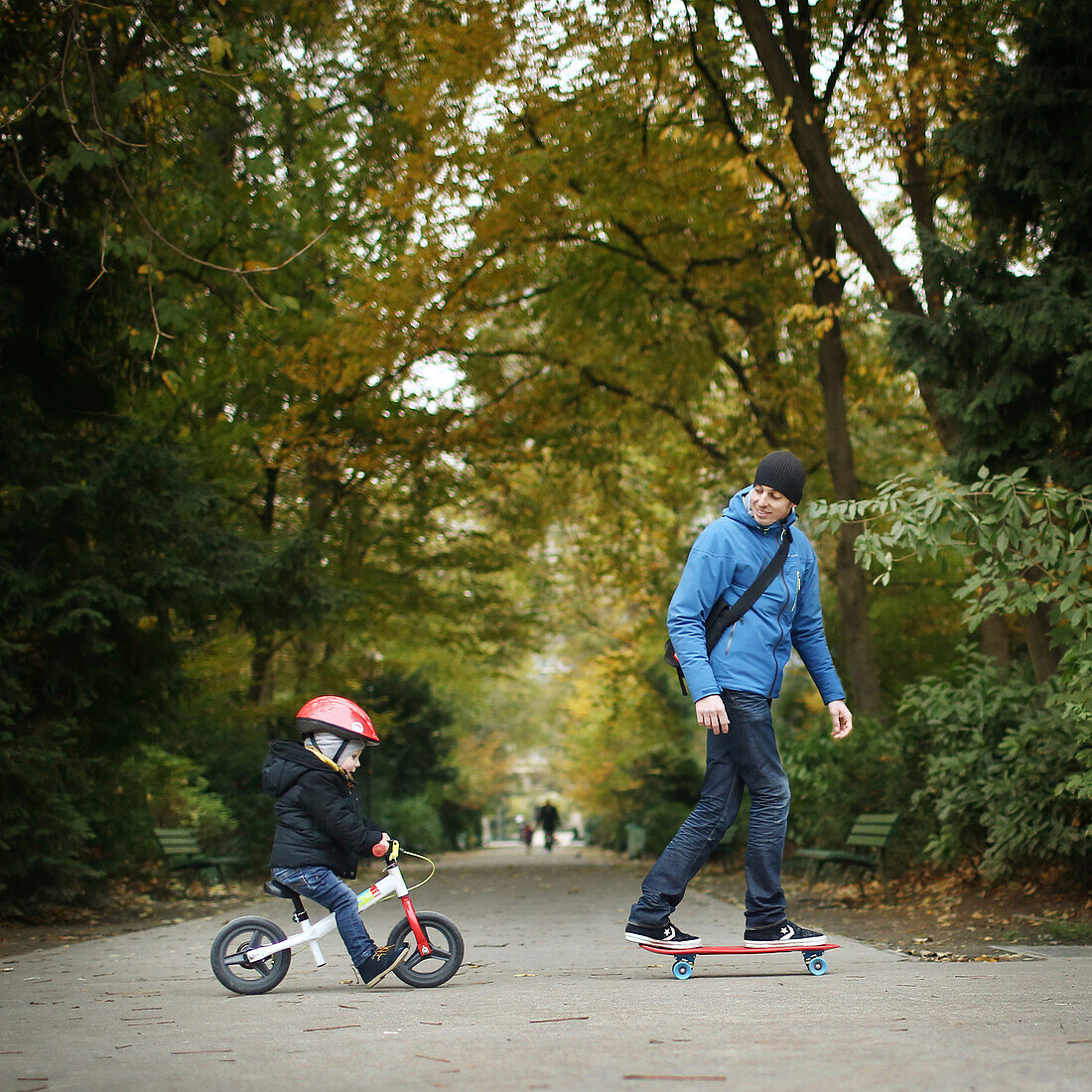 A 3 years old little boy on bike going with his father on skateboarding in a park