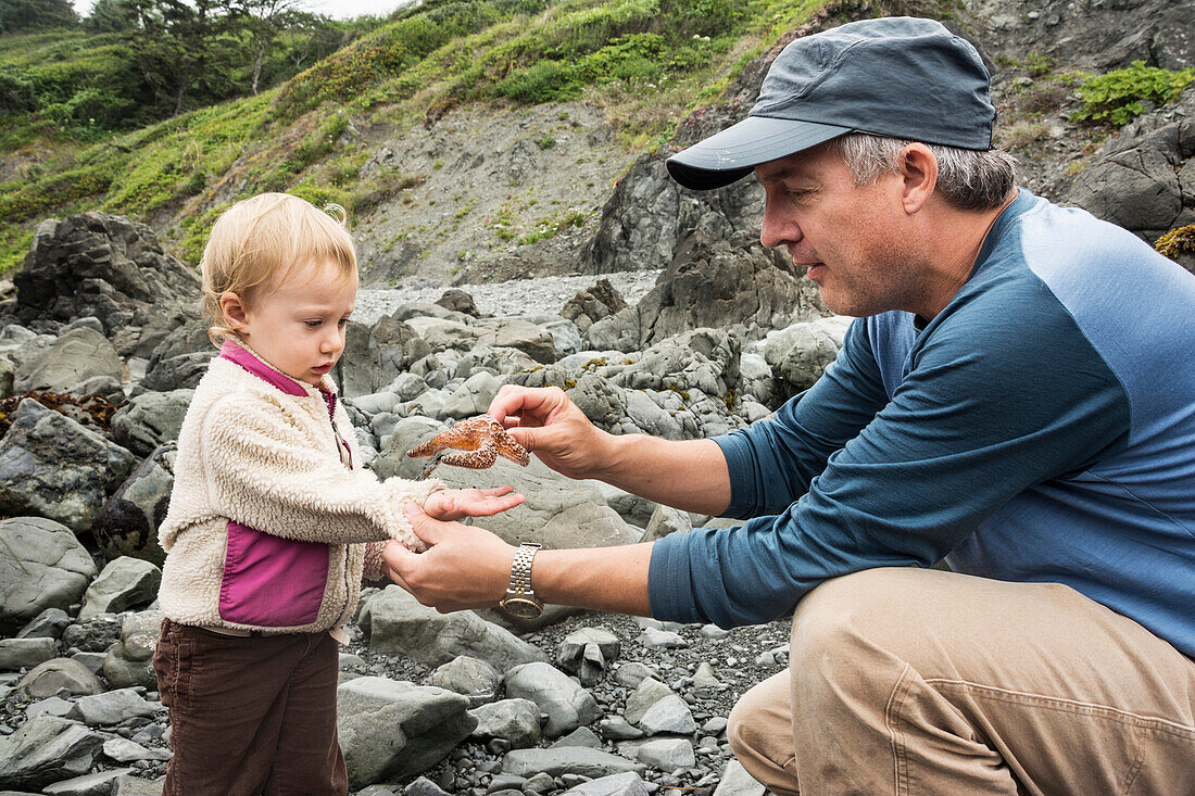 Father puts sea star in toddler girl's hand near rocky tidepools at Patrick's Point State Park, California.
