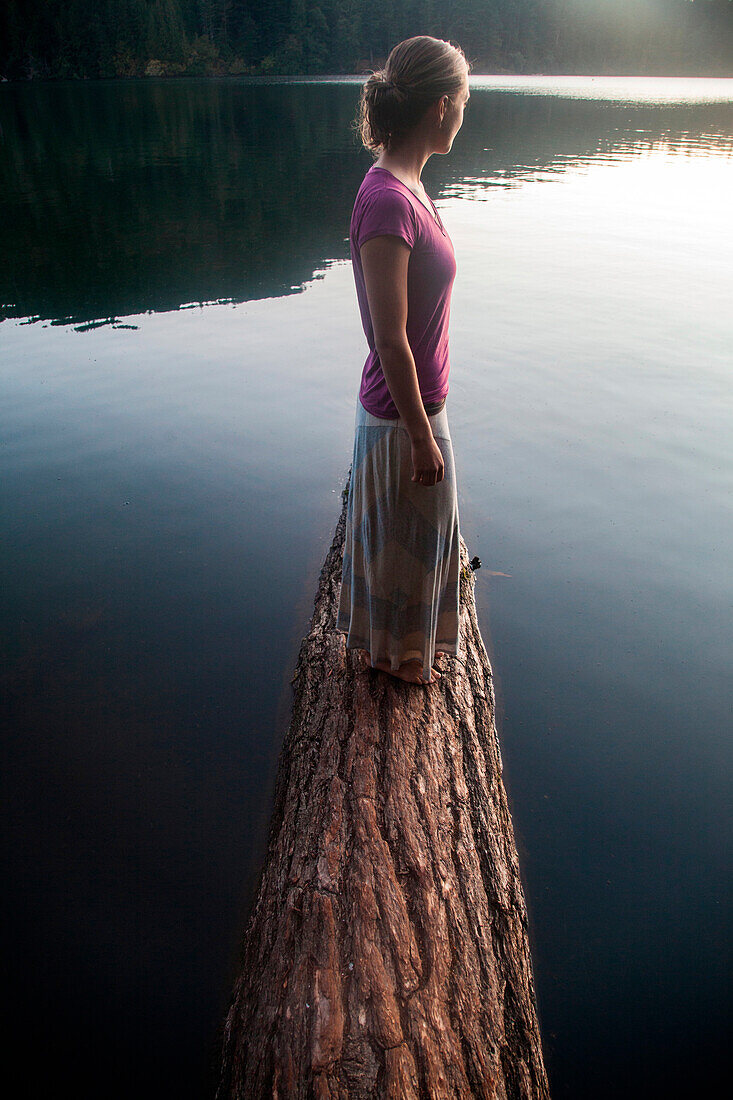 A young woman in long skirt stands on a fir tree log protruding into calm lake at sunset.