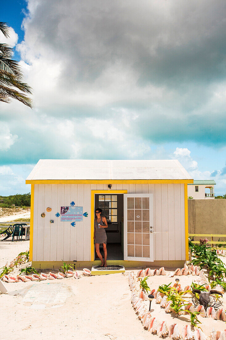 A young woman leans in the doorway of a small beachside shop surrounded by white sand.