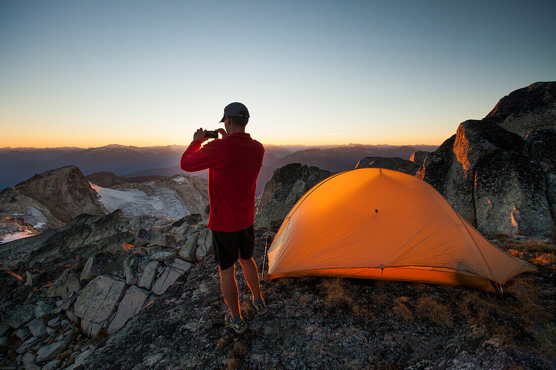 A hiker takes a picture of the sunset with his smartphone while camping on the summit of Saxifrage Peak, Pemberton, BC, Canada.
