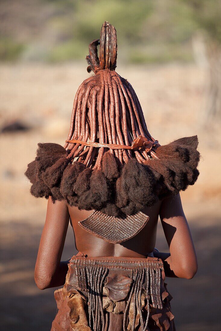 Rear view of young Himba woman showing traditional leather clothing and jewellery, hair braiding and skin covered in Otjize, a mixture of butterfat and ochre, Kunene Region (formerly Kaokoland) in the far north of Namibia