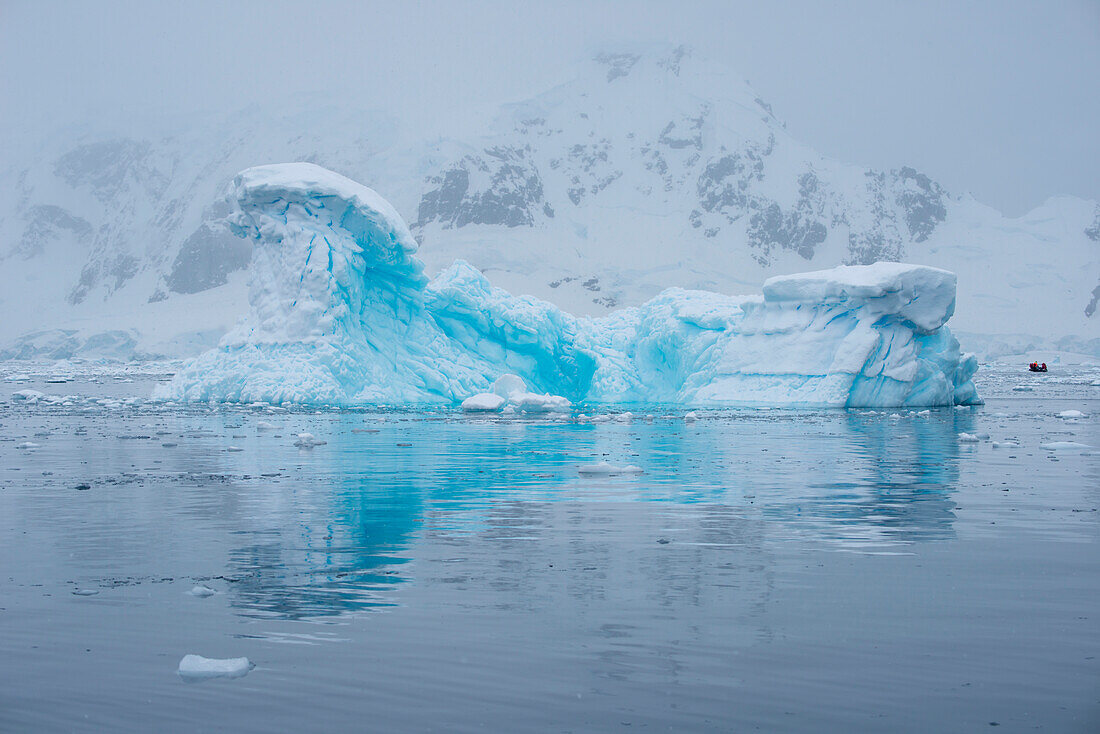A blue iceberg is reflected in the water with a Zodiac dinghy from expedition cruise ship MS Hanseatic (Hapag-Lloyd Cruises) in distance, Paradise Bay (Paradise Harbor), Danco Coast, Graham Land, Antarctica