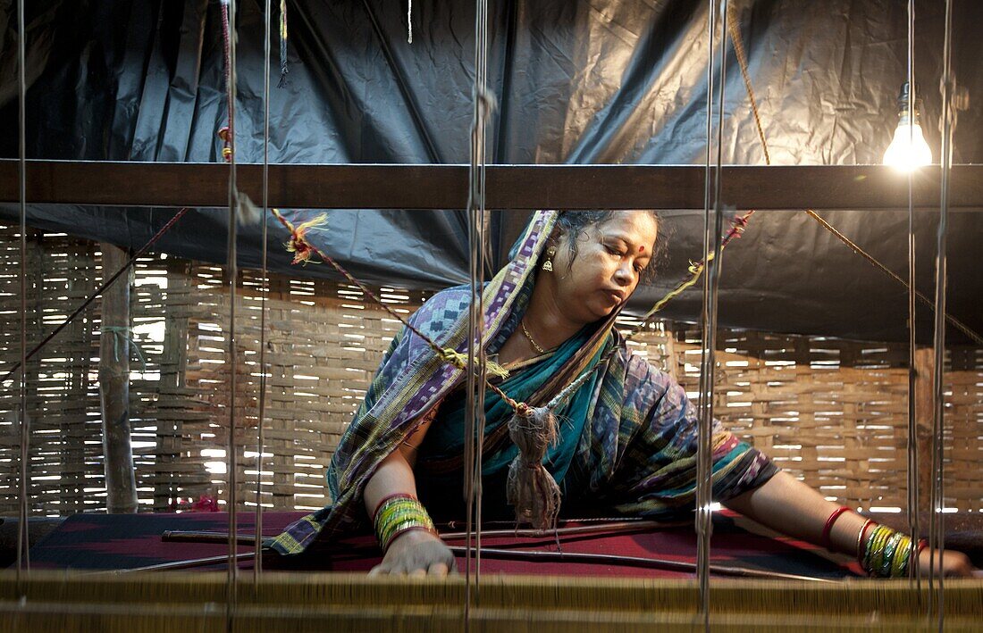 Woman in blue patterned sari weaving at loom in rough village shack, Naupatana weaving village, rural Orissa, India, Asia