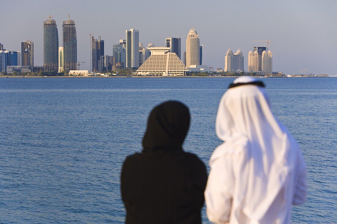 Man and woman in traditional dress looking across Doha Bay from the Corniche to the new city skyline and West Bay business and financial district, Doha, Qatar, Middle East