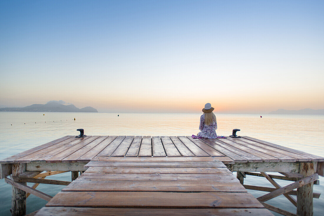 Junge blondhaarige Frau sitzt am Ende eines langen Steg in der Morgenstimmung und genießt den Ausblick auf den Sonnenaufgang. Strand Playa de Muro, Alcudia, Mallorca, Balearen, Spanien