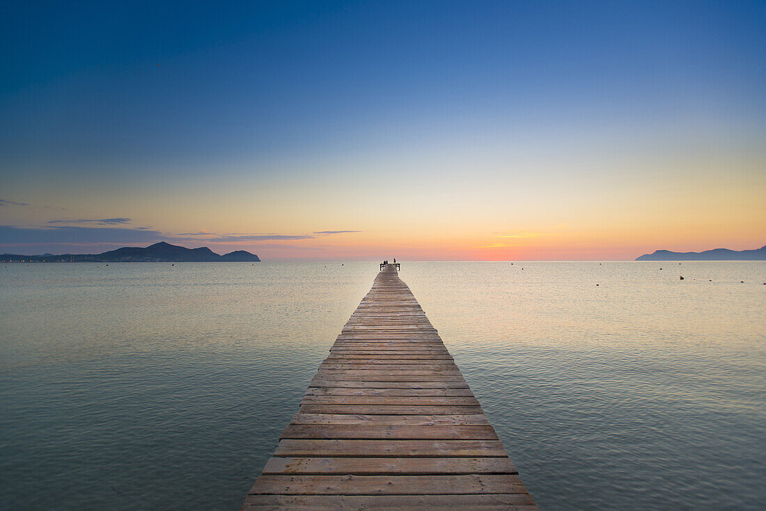 Pärchen sitzt am Ende eines langen Steg in der Morgenstimmung. Strand Playa de Muro, Alcudia, Mallorca, Balearen, Spanien