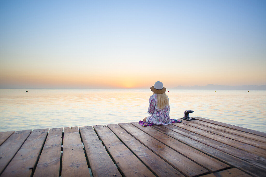 Junge blondhaarige Frau sitzt am Ende eines langen Steg in der Morgenstimmung und genießt den Ausblick auf den Sonnenaufgang. Strand Playa de Muro, Alcudia, Mallorca, Balearen, Spanien