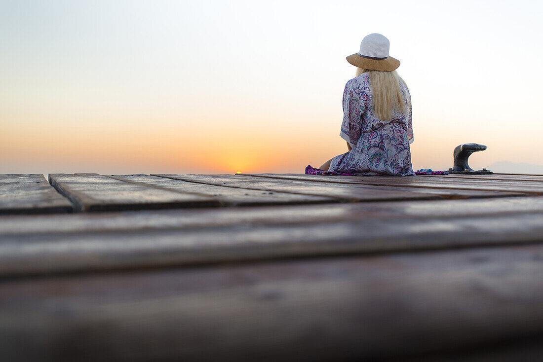 Junge blondhaarige Frau sitzt am Ende eines langen Steg in der Morgenstimmung und genießt den Ausblick auf den Sonnenaufgang. Strand Playa de Muro, Alcudia, Mallorca, Balearen, Spanien