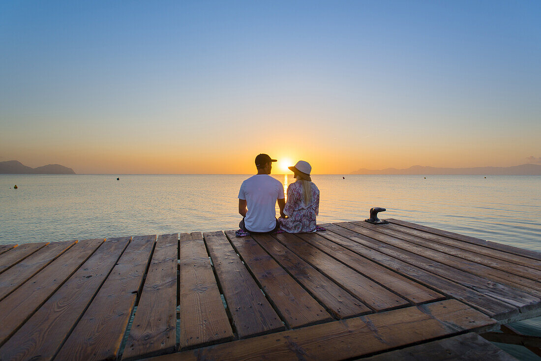 Young couple sitting at the end of a jetty in the morning atmosphere and enjoying the view of the sunrise. Playa de Muro beach, Alcudia, Mallorca, Balearic Islands, Spain