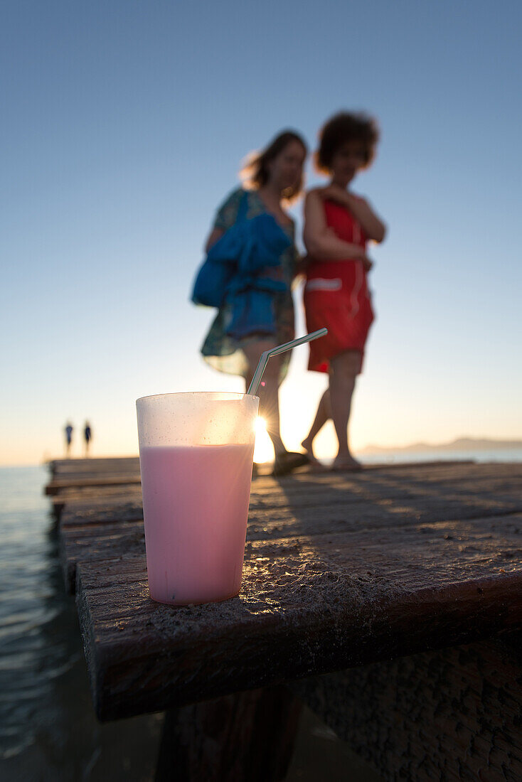 Becher mit Getränk steht auf einem Holzsteg. Im Hintergrund gehen zwei Frauen spazieren, Strand Playa de Muro, Alcudia, Mallorca, Balearen, Spanien