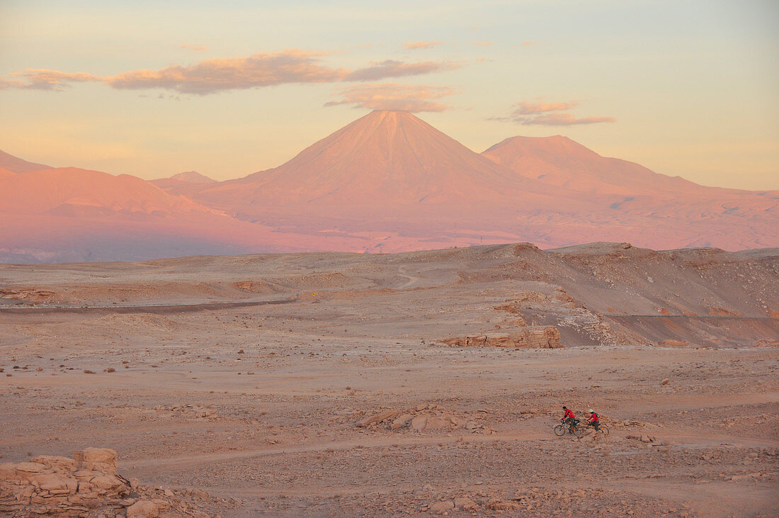 Young couple bicycling through desert, volcano Licancabur at sunset, Valle de la Luna, Valley of the moon, Atacama desert, National Reserve, Reserva Nacional Los Flamencos, Region de Antofagasta, Andes, Chile, South America