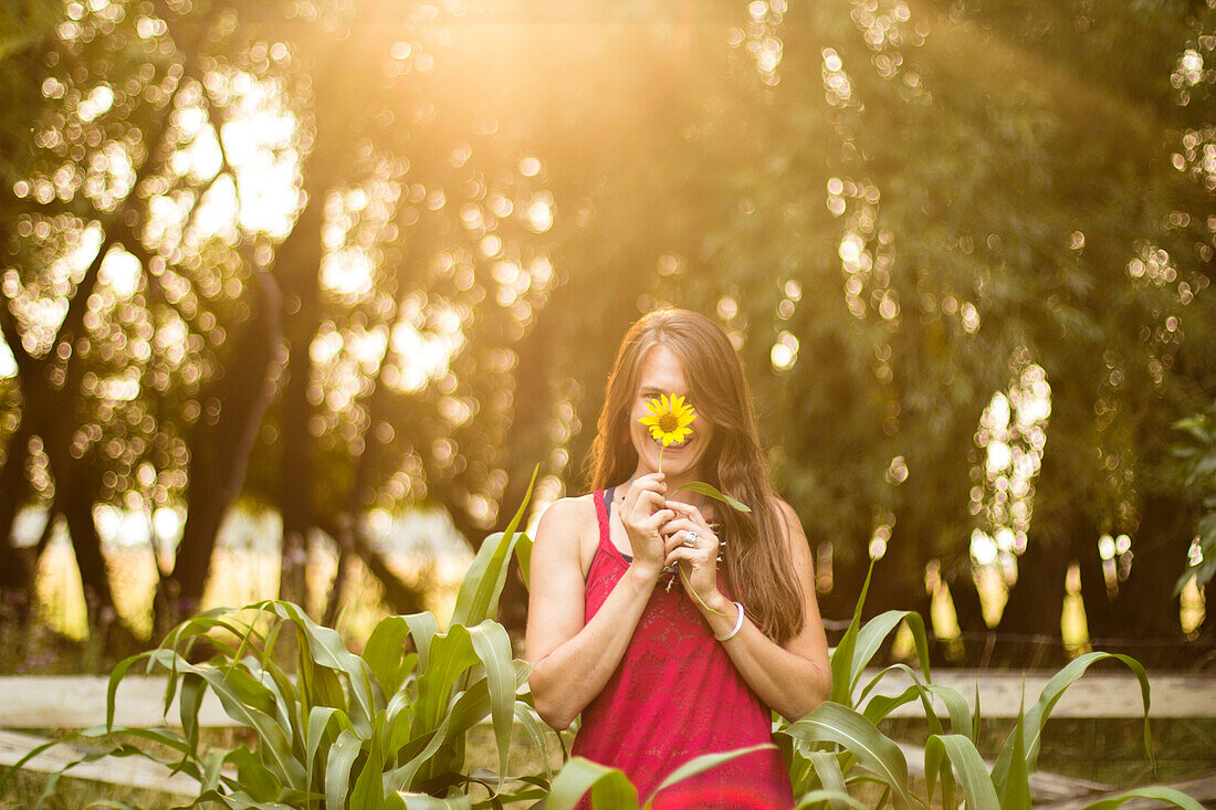 Woman smiles behind a sunflower in her garden during golden hour