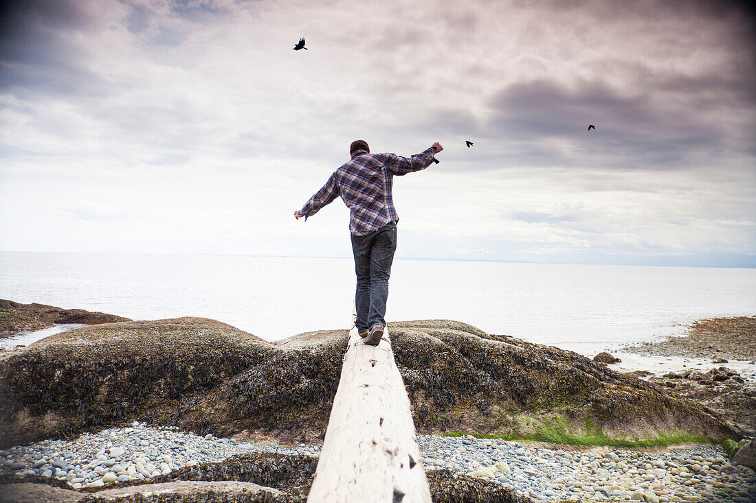 A young man balances on a log facing the Pacific Ocean at Robert's Creek Park, BC, Canada.