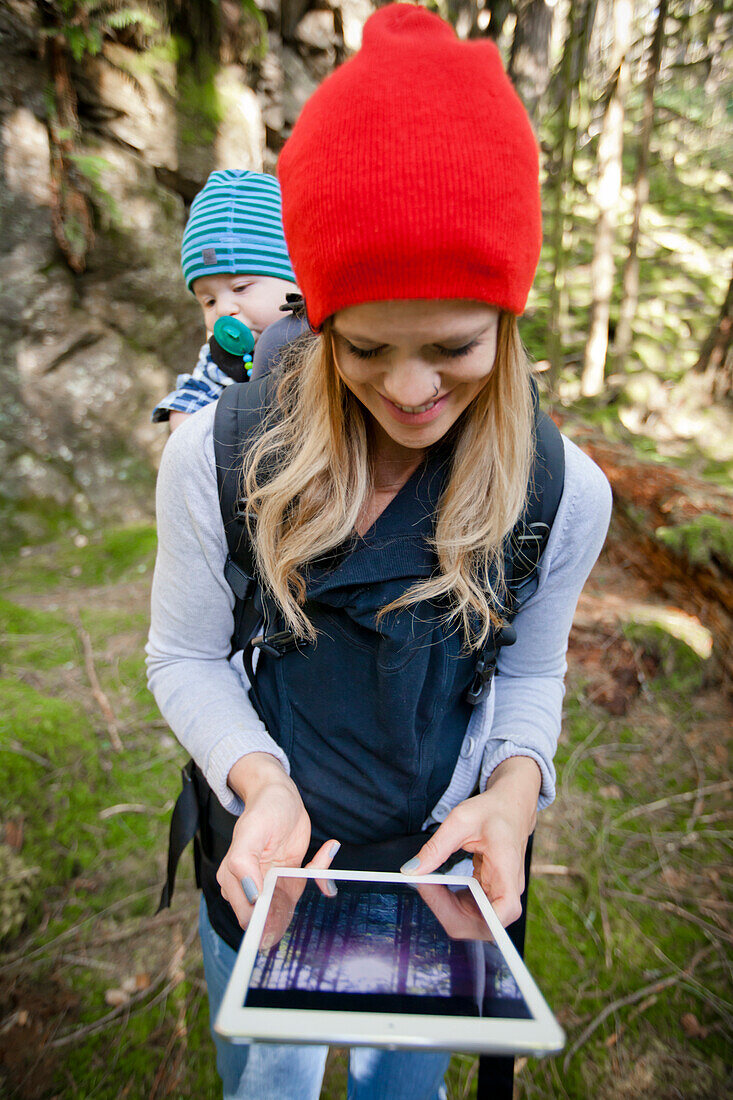 A young mother and her baby look down at a picture on a tablet in a forest setting.