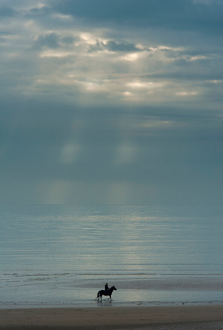 Person riding horse along Camber Sands on cloudy day with rays of sunshine, East Sussex, England