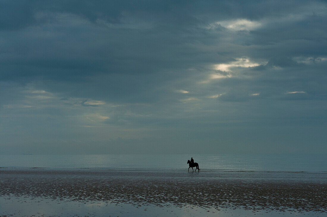 Person riding horse along Camber Sands on cloudy day with ray of sunshine, East Sussex, England