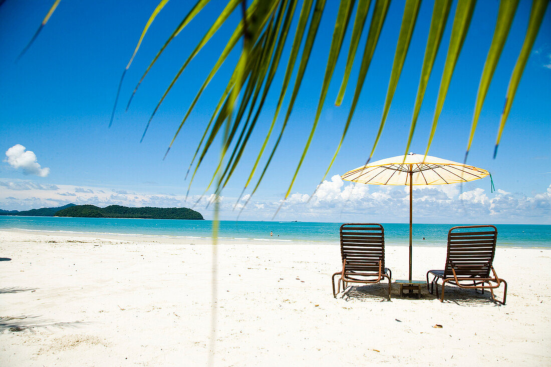 Deck chairs under parasol on white sandy beach with palm trees overlooking blue sea, Cenang beach, Pulau Langkawi, Malaysia