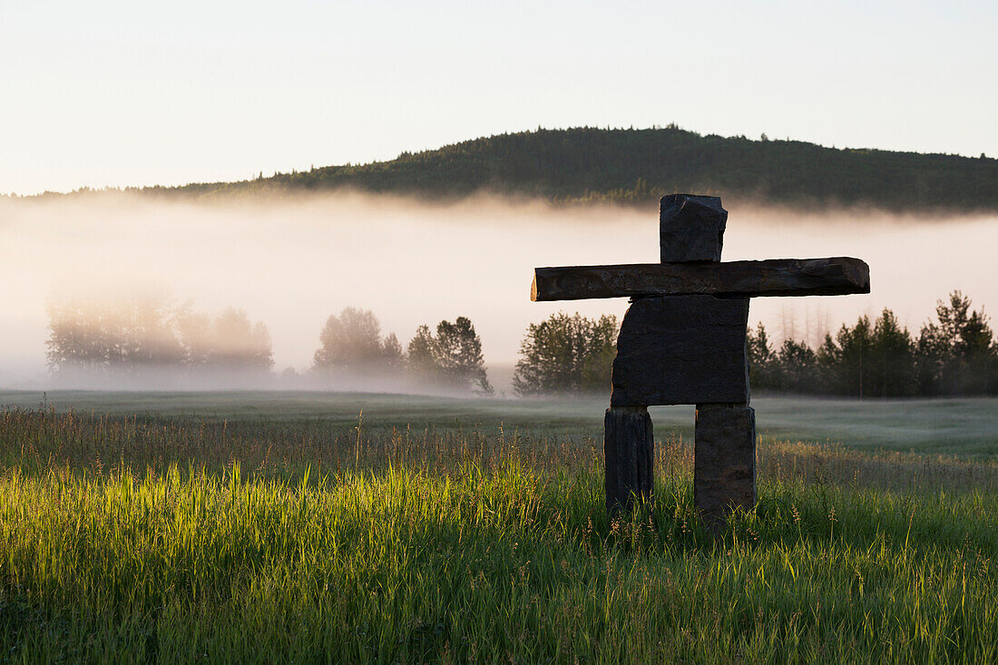 'Large rock Inuit inukshuk in grassy field with morning fog in the distance and trees with rolling hills; Alberta, Canada'