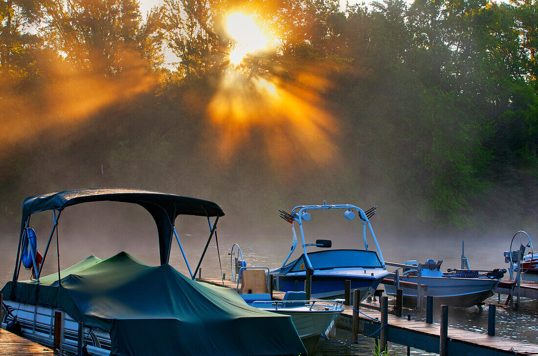 'Golden sunlight streaming through the trees to the misty lake with boats; Foster, Quebec, Canada'