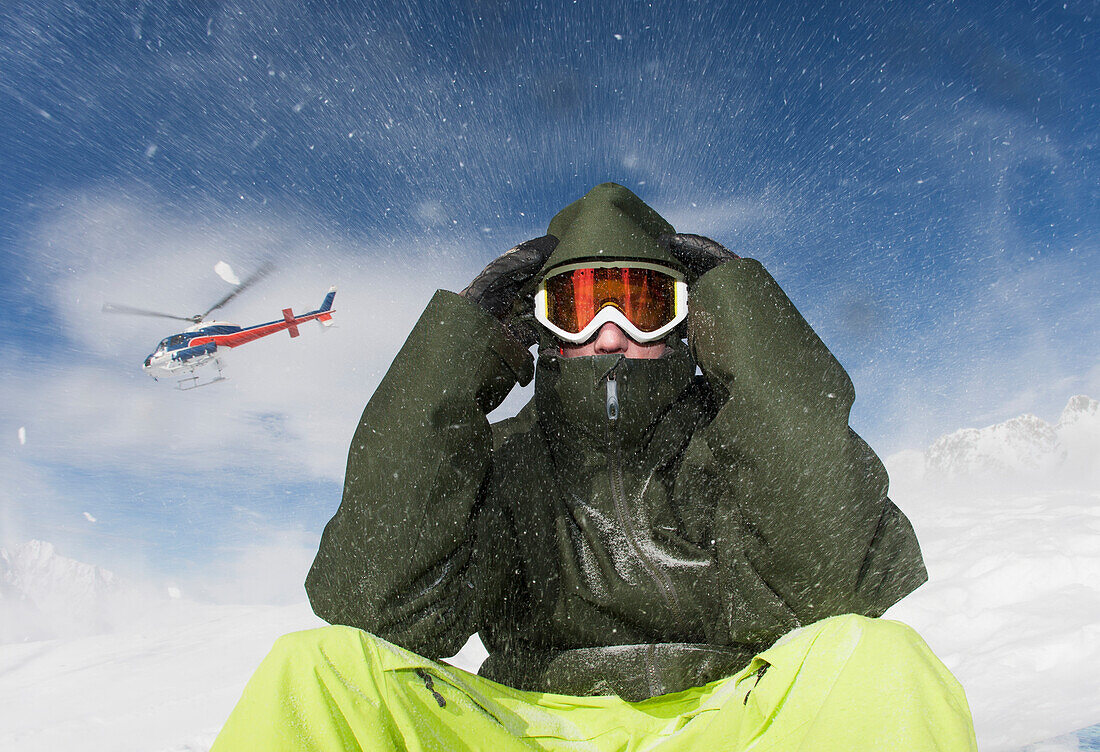 'A young man sits in winter apparel and a ski mask while a helicopter flies overhead and blows snow; Methven, New Zealand'