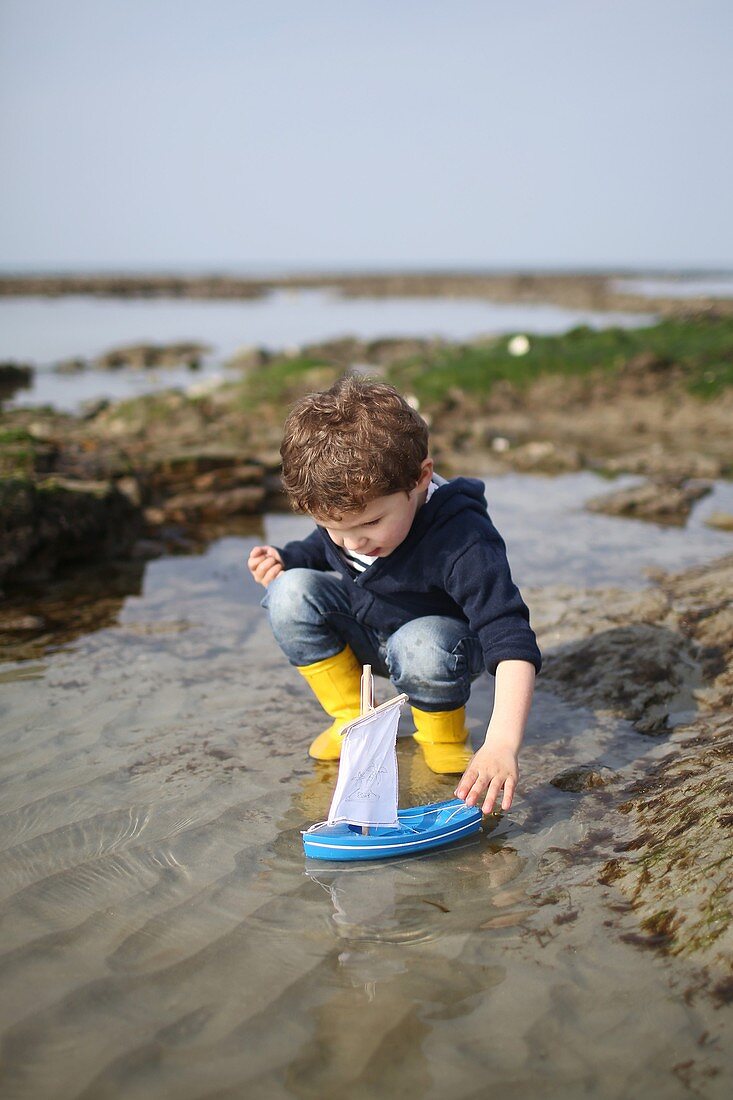 A 3 years old boy plays with a wooden boat on the beach