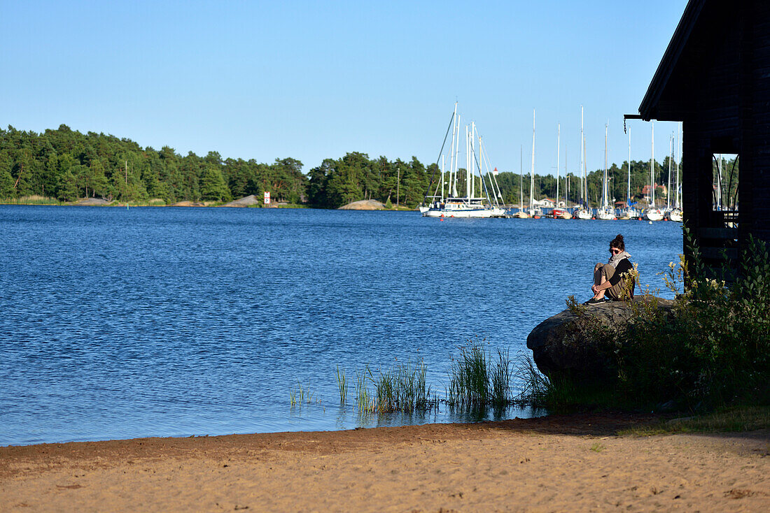 Woman sitting on a rock at the beach, with sailing boats in the background, camping ground, Vaestervik, Smaland, Sweden