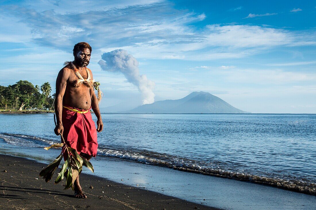 Mann läuft in traditioneller Kleidung und Schmuck am schwarzen Sandstrand von Rabaul im Norden Papua Neuguineas. Im Hintergrund der aktive Vulkan Tavurvur (Hornissennest) auf Matupi Island, Papua Neuguinea, Neu Britannien, Süd Pazifik