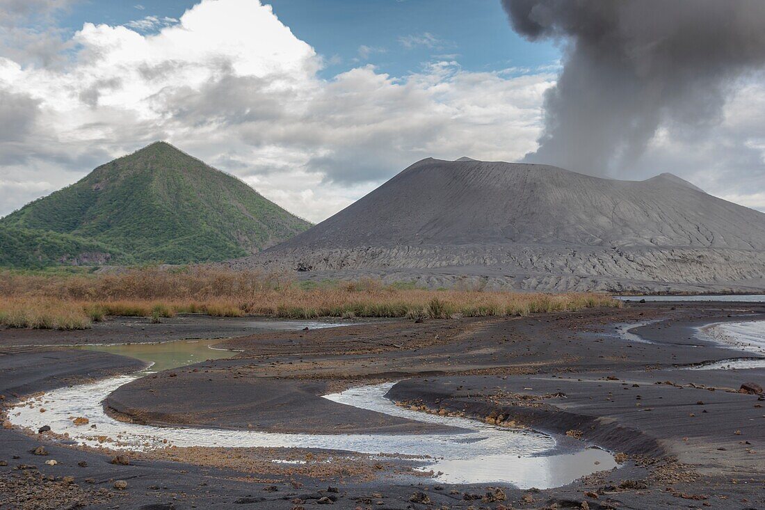 Eruption of the active volcano Tavurvur with ash cloud. In the foreground rainwater in the ash sand, in the background the green cone of the „Vulcan'' volcano, Papua New Guinea, New Britain, South Pacific