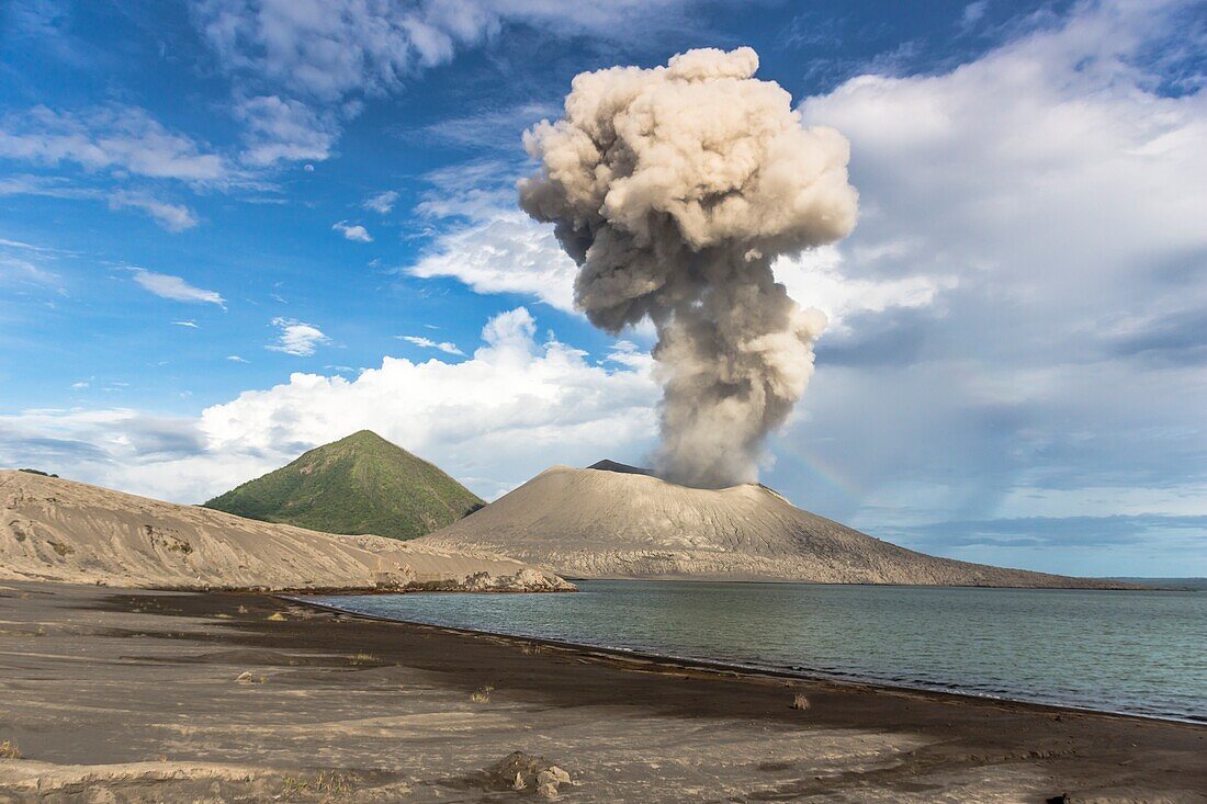 Eruption of the active volcano Tavurvur with ash cloud and rainbow; ash beach and ocean surf in the front; the green cone apex of the Vulcan in the background,  Papua New Guinea, New Britain, South Pacific