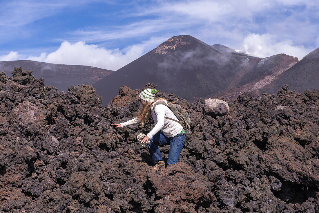 Solidified lava field of the Etna volcano with lava river at SE crater in the background. Woman running over solidified and sharp-edged lava, Sicily, Italy