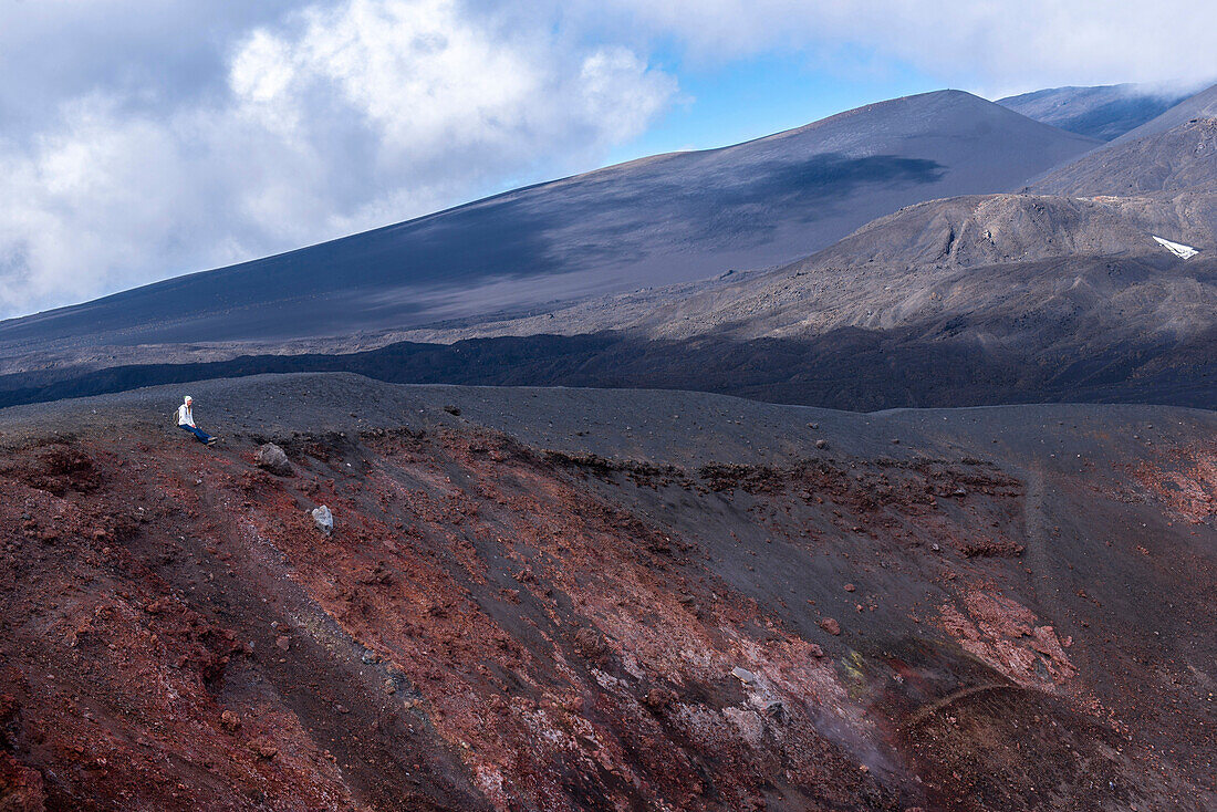 Woman sitting at the edge of an extinct crater at the foot of the Etna volcano. Minerals in the soil are the reason for the red color of the rocks, Sicily, Italy