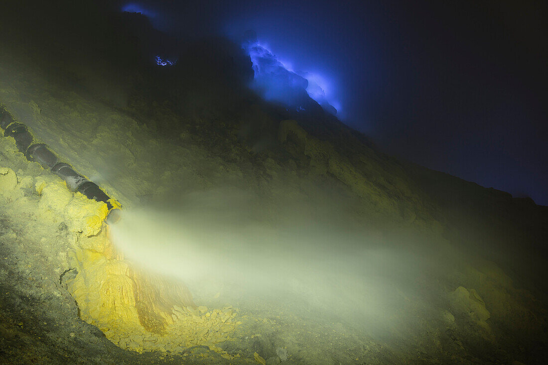 Burning sulfur and blue flames of the volcano Ijen at night. Pipe transferring hot sulfur gases for cooling down. Sulfur depositing in solid form, East Java, Ijen volcano, Indonesia