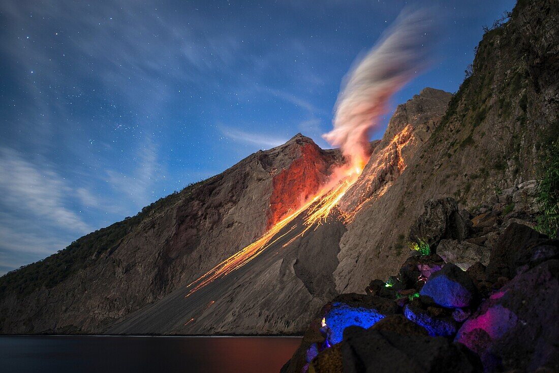 Eruption of Batu Tara volcano at full moon with the Milky Way. Ash eruptions and lava bombs flying on the fire slide towards the sea. Stones in the foreground are illuminated in different colors, island of Komba, Flores Sea, Indonesia