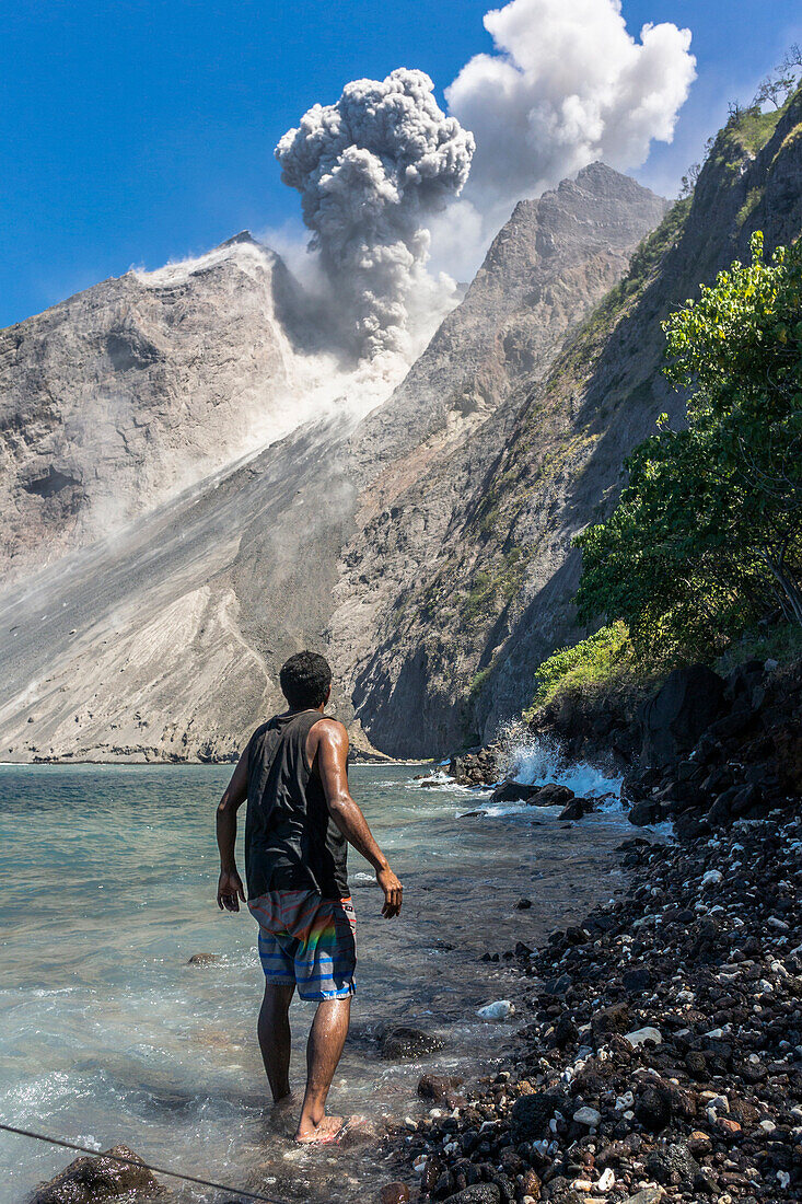 Man standing at the foot of active Batu Tara of Batu Tara volcano between the sea and cliffs and getting frightened by a large eruption. - Indonesia, island of Komba, Flores Sea