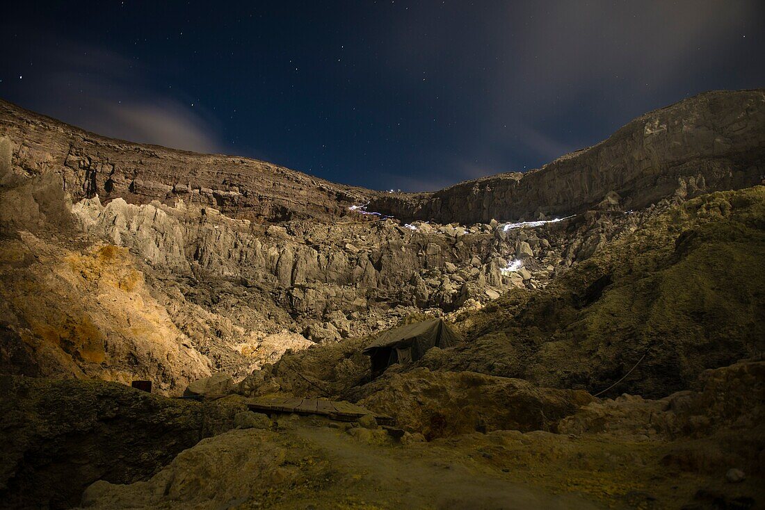 Blick vom dem Kraterboden des aktiven Vulkans Ijen auf den Kraterrand bei Nacht während des Vollmondes.  Kraterflanken werden durch den Mond ausgeleuchtet. Leuchtspuren durch Taschenlampen in der Felswand, Sterne am Himmel. - Indonesien, Ost-Java, Vulkan 