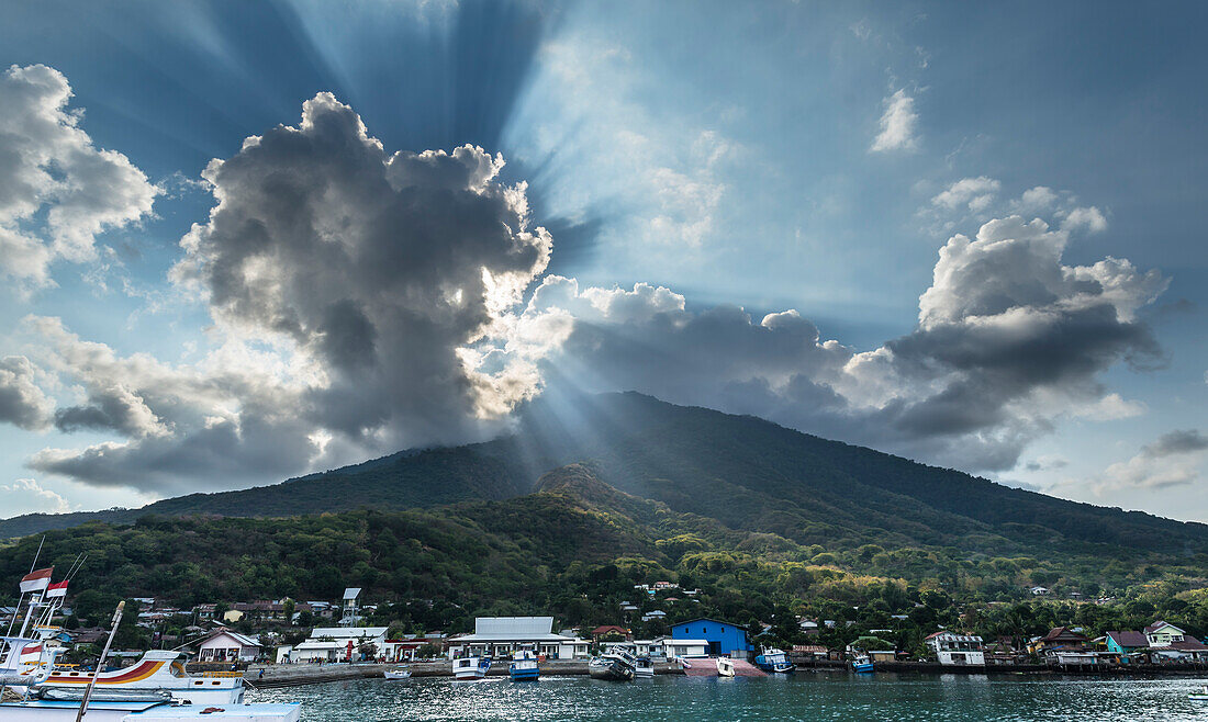 Cone of an extinct volcano, dense forests growing to the port at the coast. Sunrays reaching through the clouds and shining through the forest, Flores Island, Indonesia