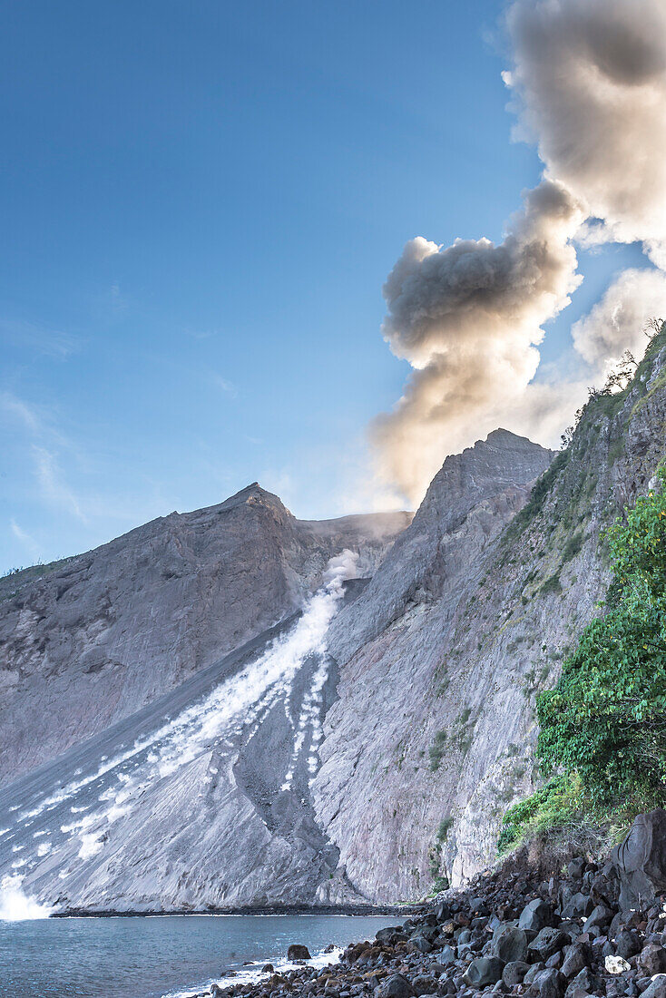 Große Asche Eruption und Gesteins Auswurf auf die Feuerflanke des Vulkans Batu Tara mit austretenden Vulkan-Gasen und Asche in der Flores Sea bei Tage vor blauem Himmel mit steiniger Küste, Meer, und Vegetation im Vordergrund. Gesteinsbrocken rollen in da