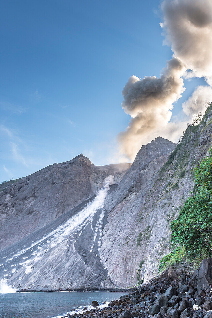 Large ash eruption with ejections of rocks at the burning edge of Batu Tara in the Flores Sea with emitting volcanic gases and ash with blue sky. Stony coast, sea, and vegetation in the foreground, Rocks rolling into the open sea, island of Komba, Flores 
