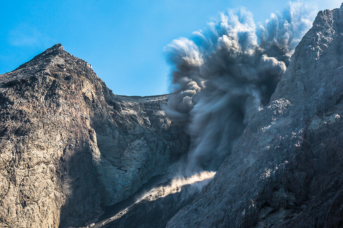 Large ash eruption with ejections of rocks at the burning edge of Batu Tara in the Flores Sea with emitting volcanic gases and ash and blue sky. Stony coast, sea, and vegetation in the foreground, Rocks rolling into the open sea, island of Komba, Flores S