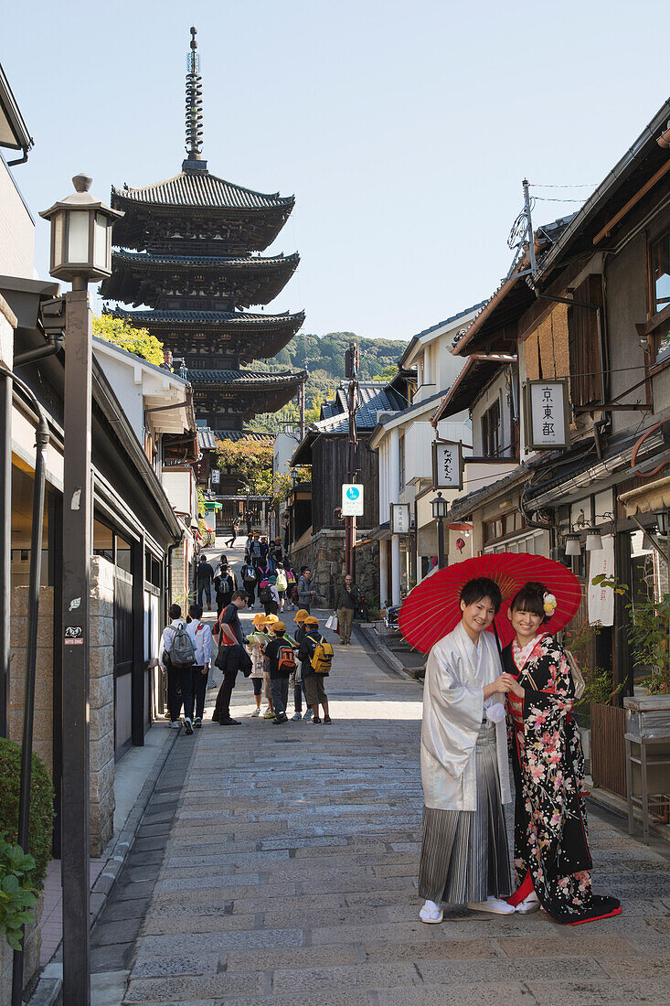 Young Japanese couple in kimono posing under a red umbrella in a narrow street with pagoda in the distance, Kyoto, Japan