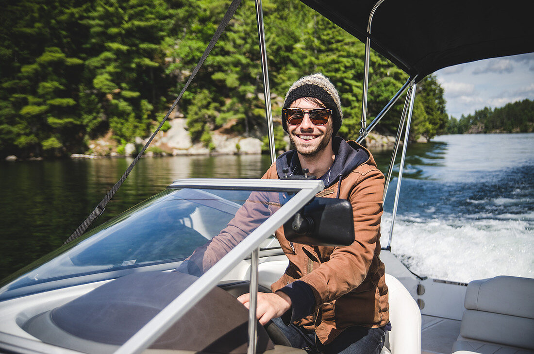 Smiling Young Adult Man in Wool hat Driving Motor Boat on Lake