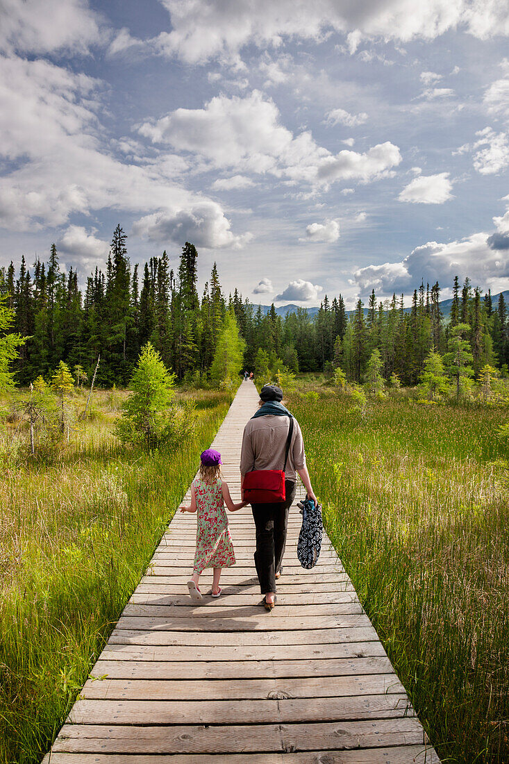 A mother and daughter walk down a boardwalk through a wetland surrounded by evergreen trees, Liard River Hot Springs Provincial Park, British Columbia, Canada, Summer