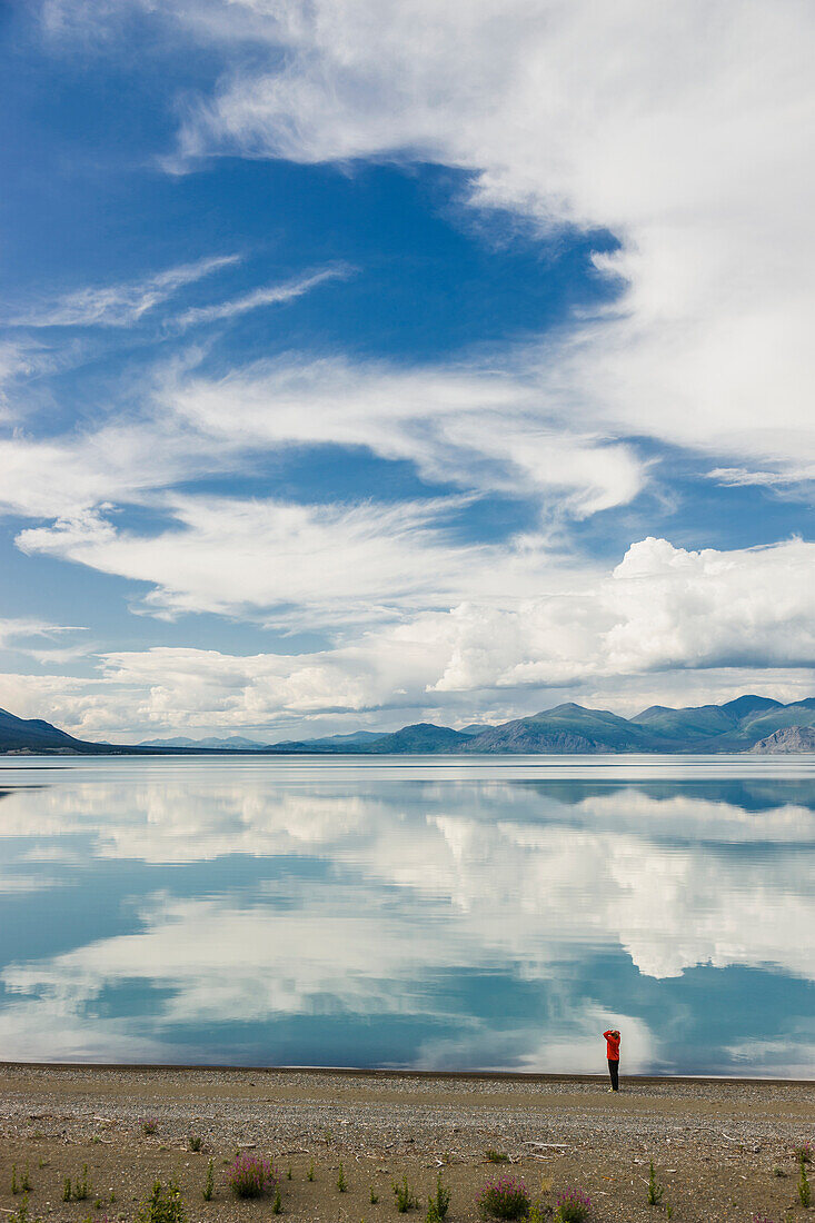 A woman stands at the shore of Kluane Lake with mountains and clouds reflecting in the calm waters of Kluane Lake, Yukon Territory, Canada, Summer