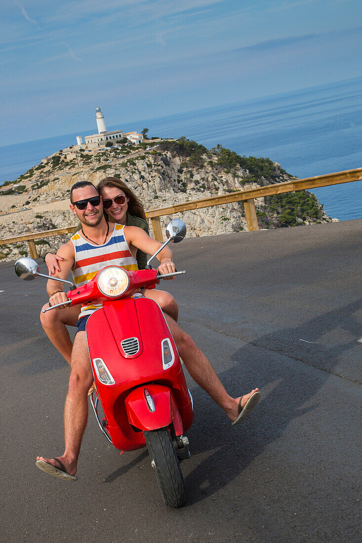 Young couple riding a red Vespa scooter on road along Cap de Formentor peninsula with Faro de Formentor lighthouse behind, Palma, Mallorca, Balearic Islands, Spain