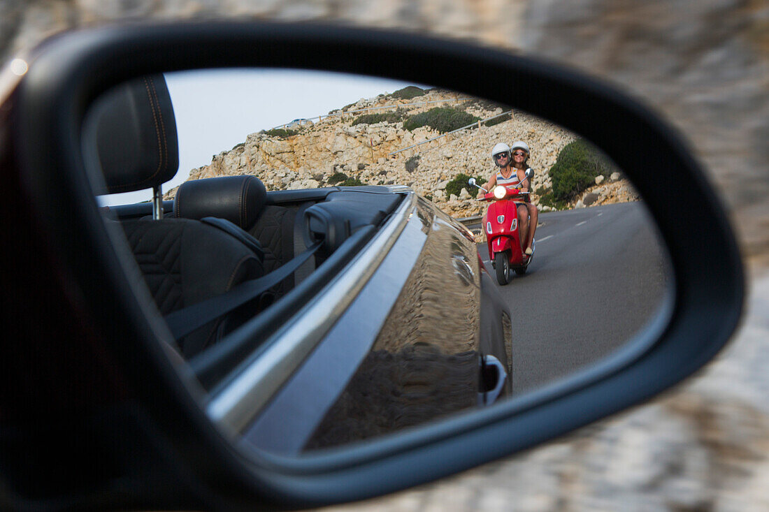 Blick in den Rückspiegel eines Sunny Cars Cabriolets auf junges Paar auf rotem Vespa Motorroller auf Straße entlang der Halbinsel Cap de Formentor, Cap de Formentor, Palma, Mallorca, Balearen, Spanien