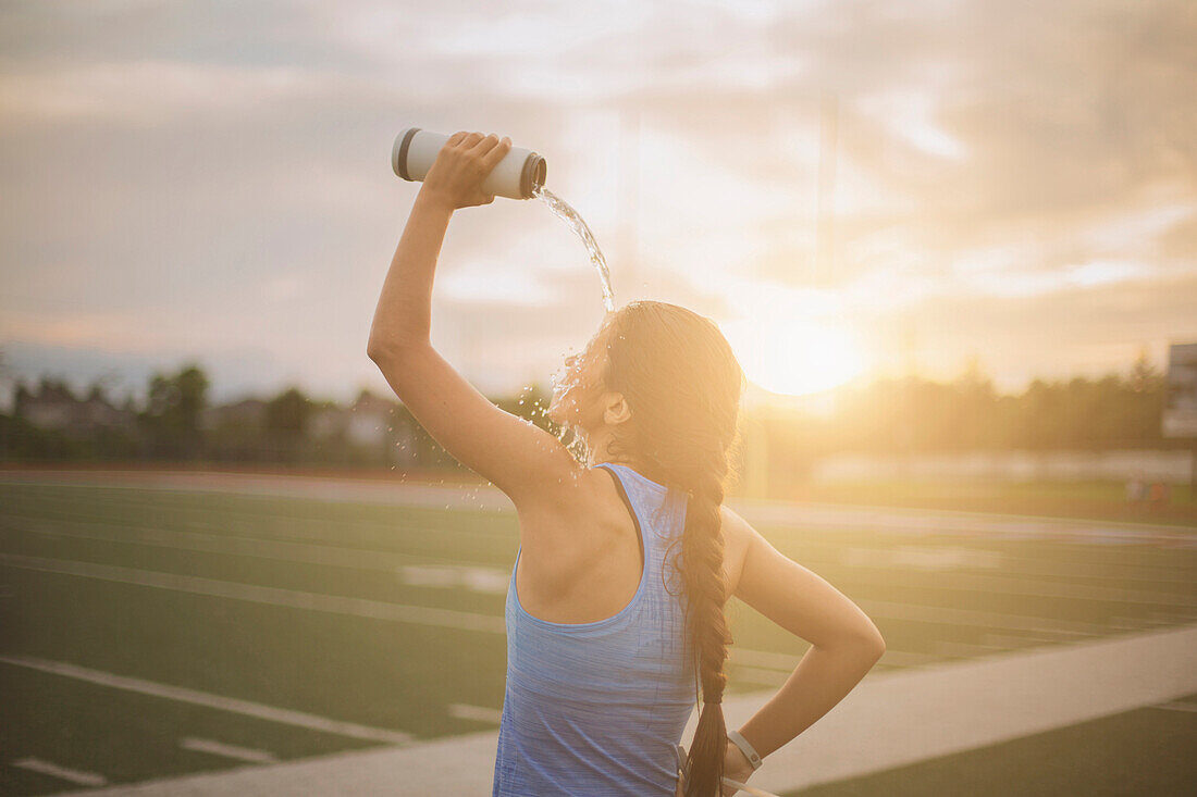 Gemischtrassige Athletin gießt sich auf einem Sportplatz eine Wasserflasche ein