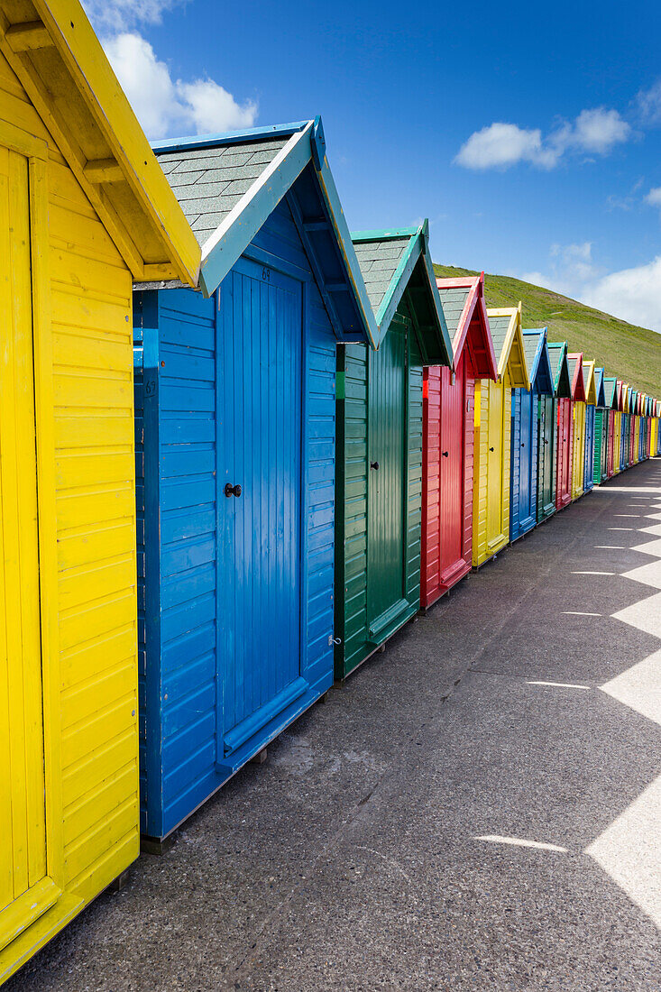 Row of colourful beach huts and their shadows with green hill backdrop, West Cliff Beach, Whitby, North Yorkshire, England, United Kingdom, Europe