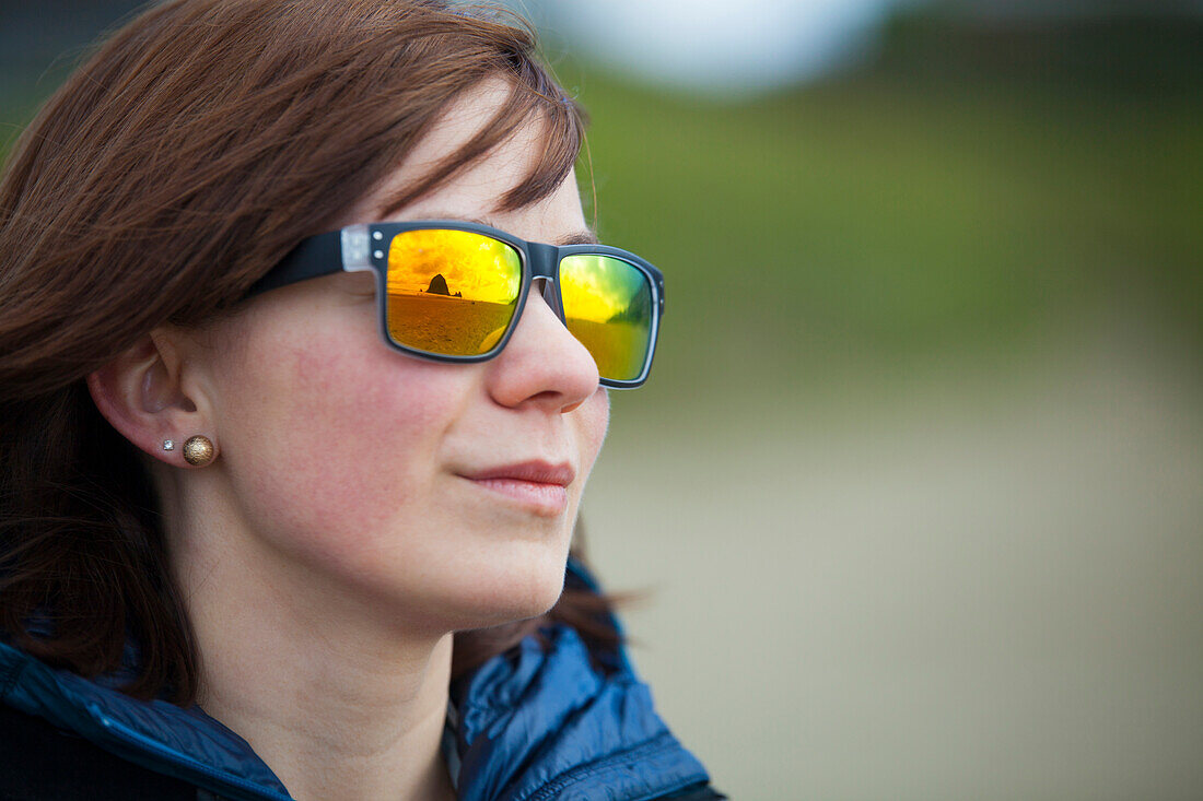 Haystack Rock and Cannon Beach is reflected in a young womans colorful sunglasses.